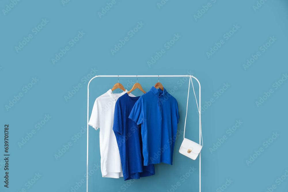 Stylish t-shirts and bag hanging on rack against color wall