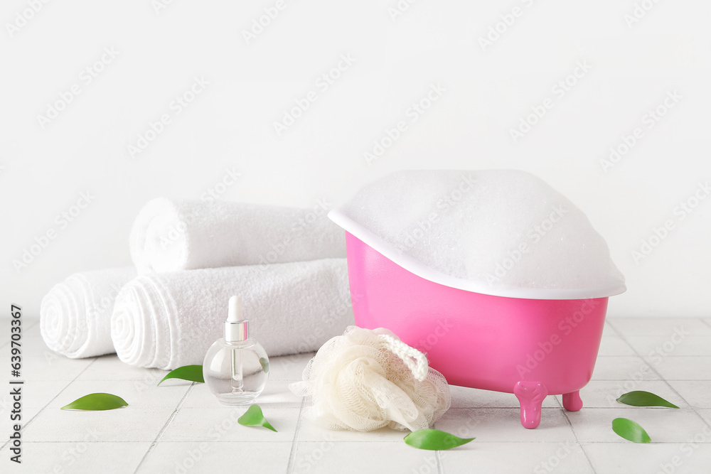 Small bathtub with foam, bath supplies and plant leaves on light tile table