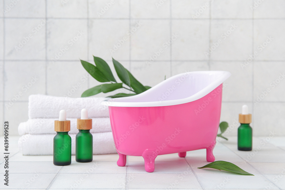 Small bathtub, clean towels, bottles of essential oil and plant branch on light tile background