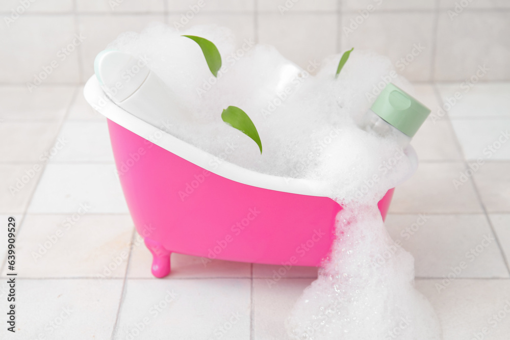 Small bathtub with foam and cosmetic products on light tile background