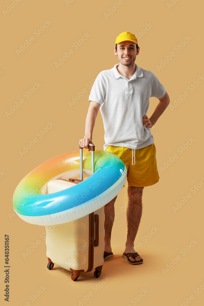 Young man with inflatable ring and suitcase on beige background