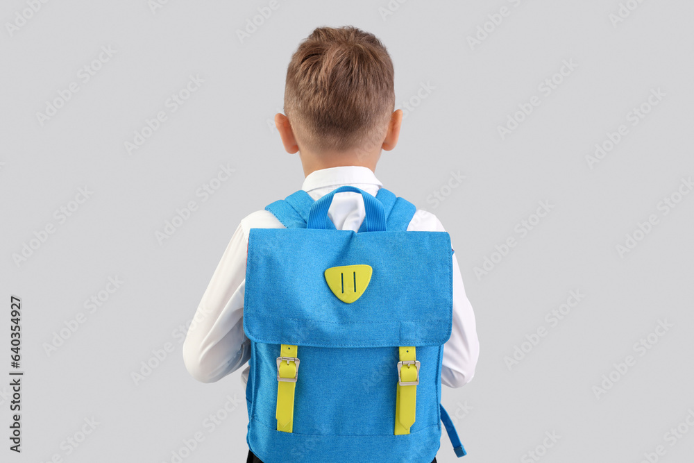 Little schoolboy with blue backpack on grey background, back view