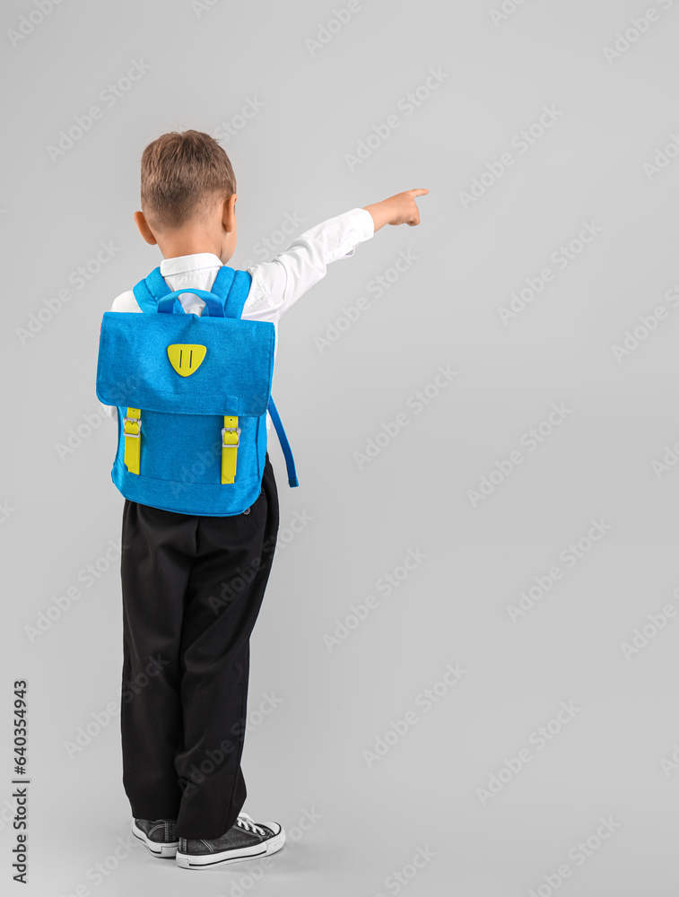 Little schoolboy with blue backpack pointing at something on grey background, back view