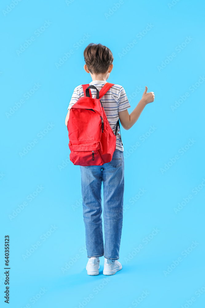 Little boy with schoolbag showing thumb-up on blue background, back view