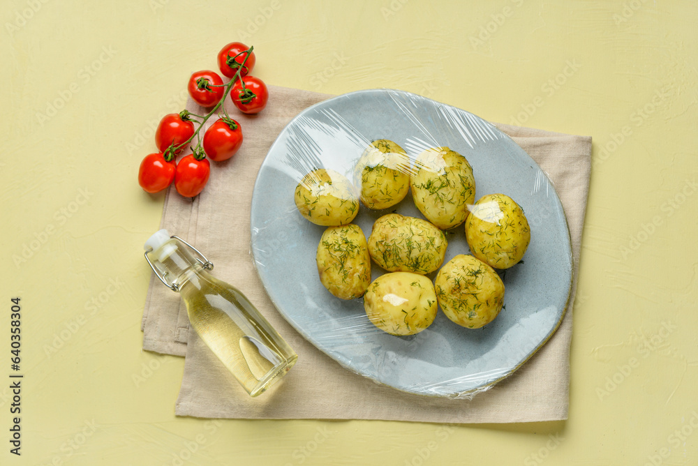 Plate of tasty boiled potatoes wrapped with stretch wrap, tomatoes and oil on color background