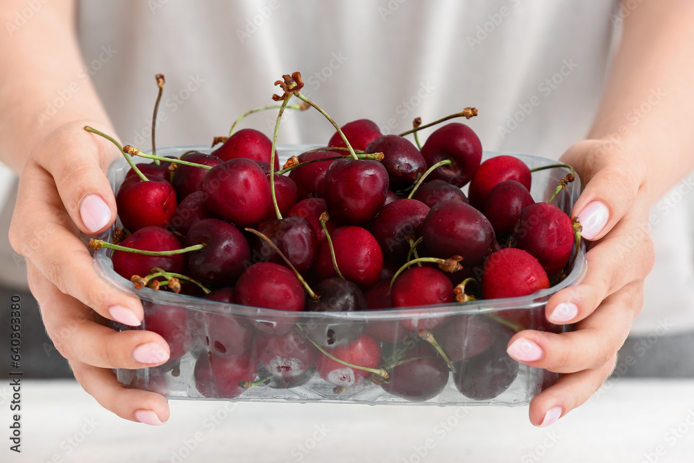 Woman holding plastic container with ripe cherries at table in kitchen