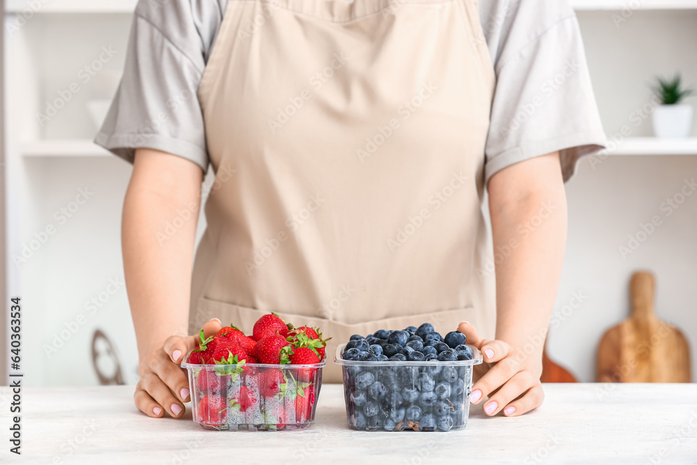 Woman holding plastic containers with ripe berries at table in kitchen