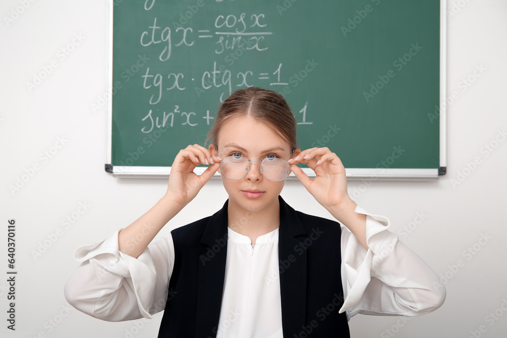 Young math teacher with eyeglasses sitting at wooden table in classroom