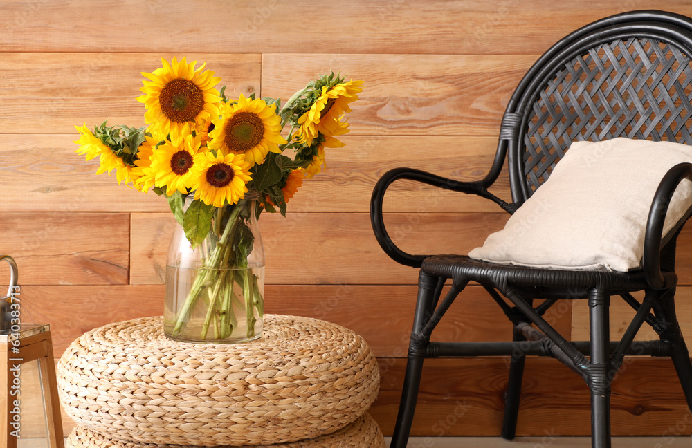 Vase with beautiful sunflowers on wicker pouf and armchair near wooden wall, closeup