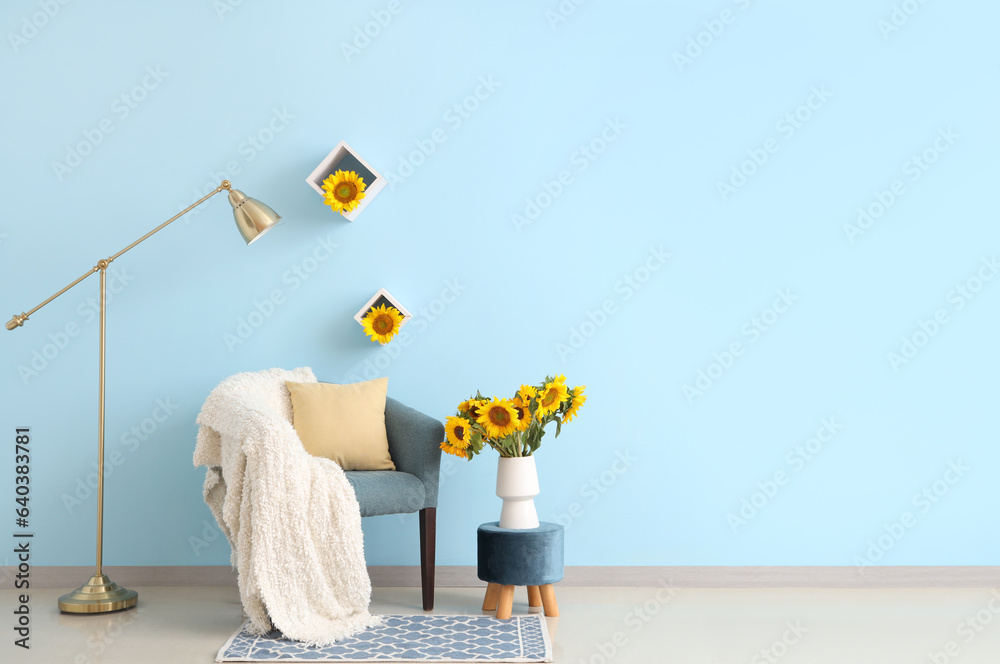 Vase with beautiful sunflowers on pouf and cozy armchair near blue wall