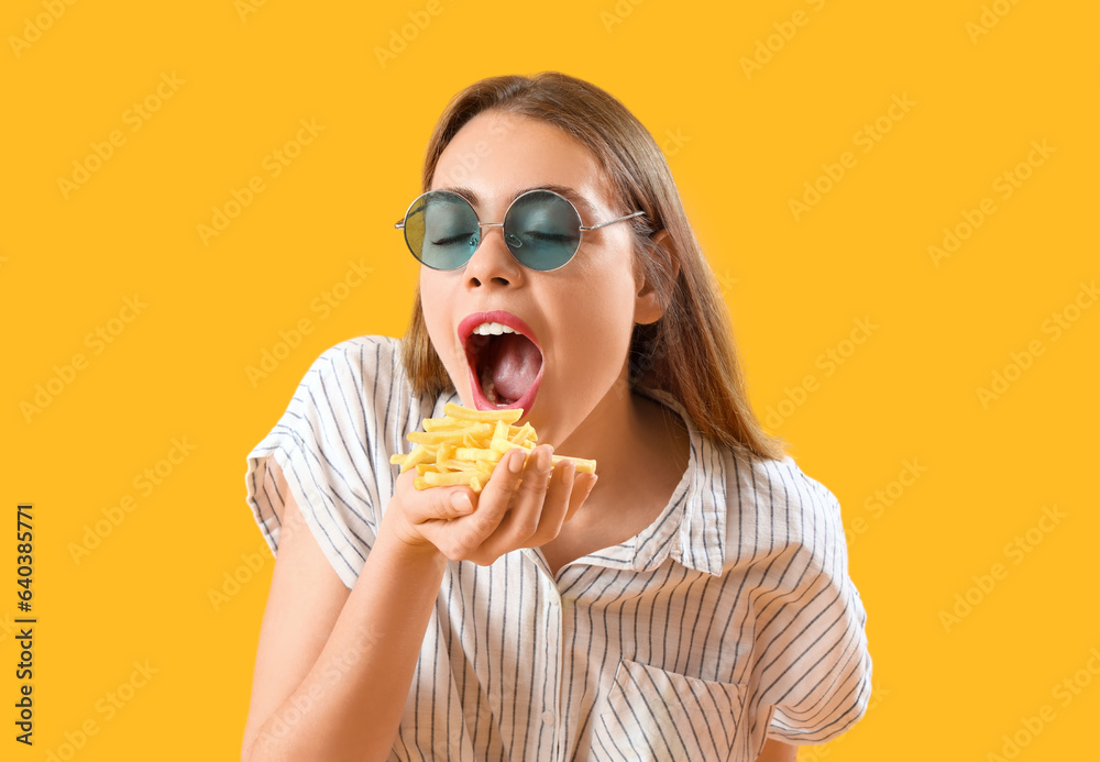 Beautiful young woman in sunglasses eating french fries on yellow background