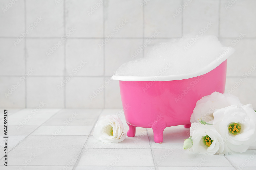 Small bathtub with foam and beautiful eustoma flowers on light tile background