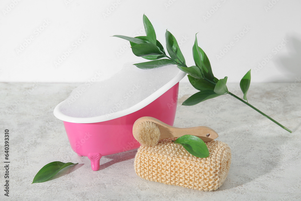 Small bathtub with soap foam, massage brush, sponge and plant branch on table against light backgrou