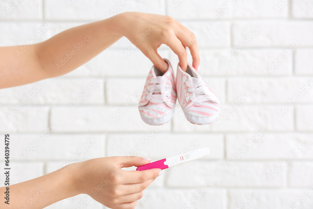 Female hands with positive pregnancy test and baby booties against light brick wall, closeup