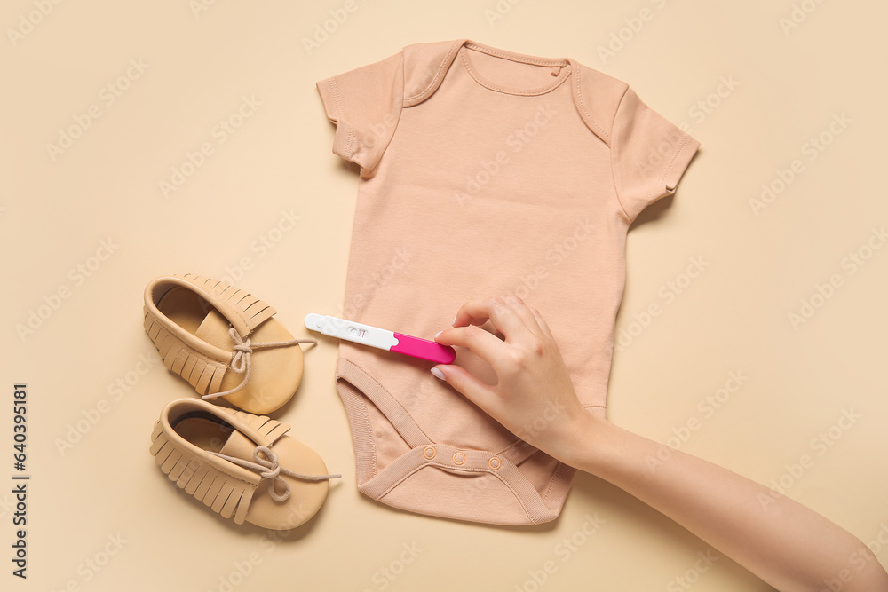 Female hand with pregnancy test, baby booties and bodysuit on color background