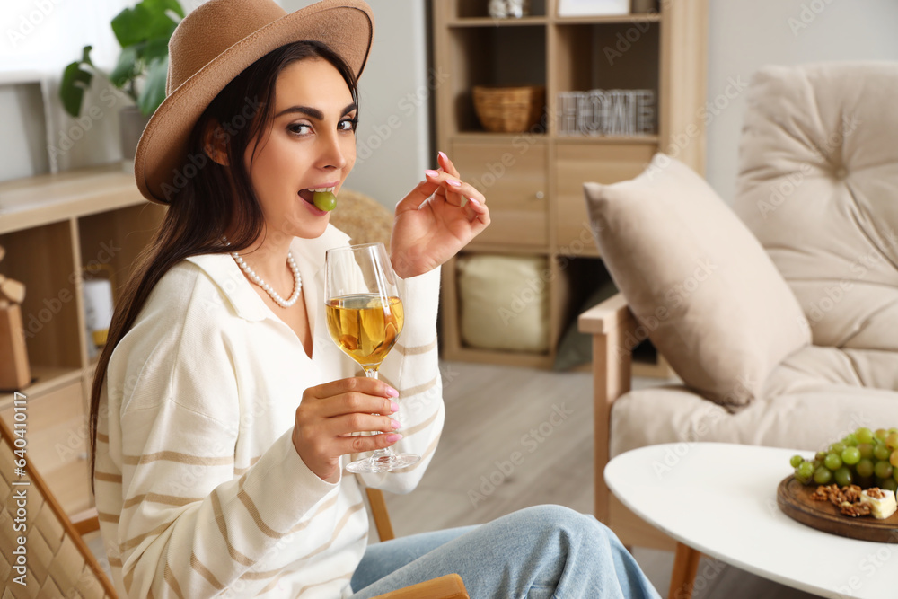 Young woman with glass of wine eating grape in armchair at home