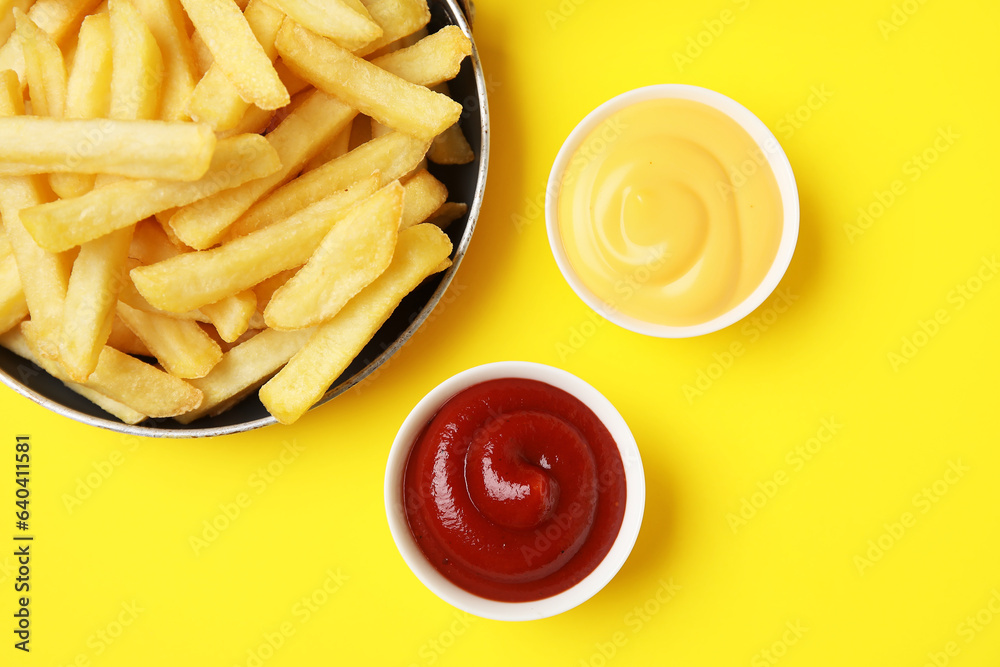 Frying pan with golden french fries, ketchup and cheese sauce on yellow background, closeup