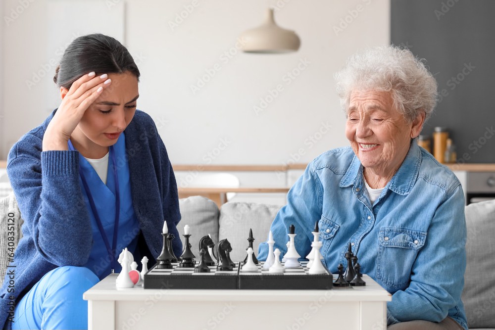 Young caregiver with senior woman playing chess at home