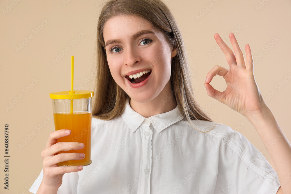 Young woman with glass of carrot juice showing OK on beige background, closeup