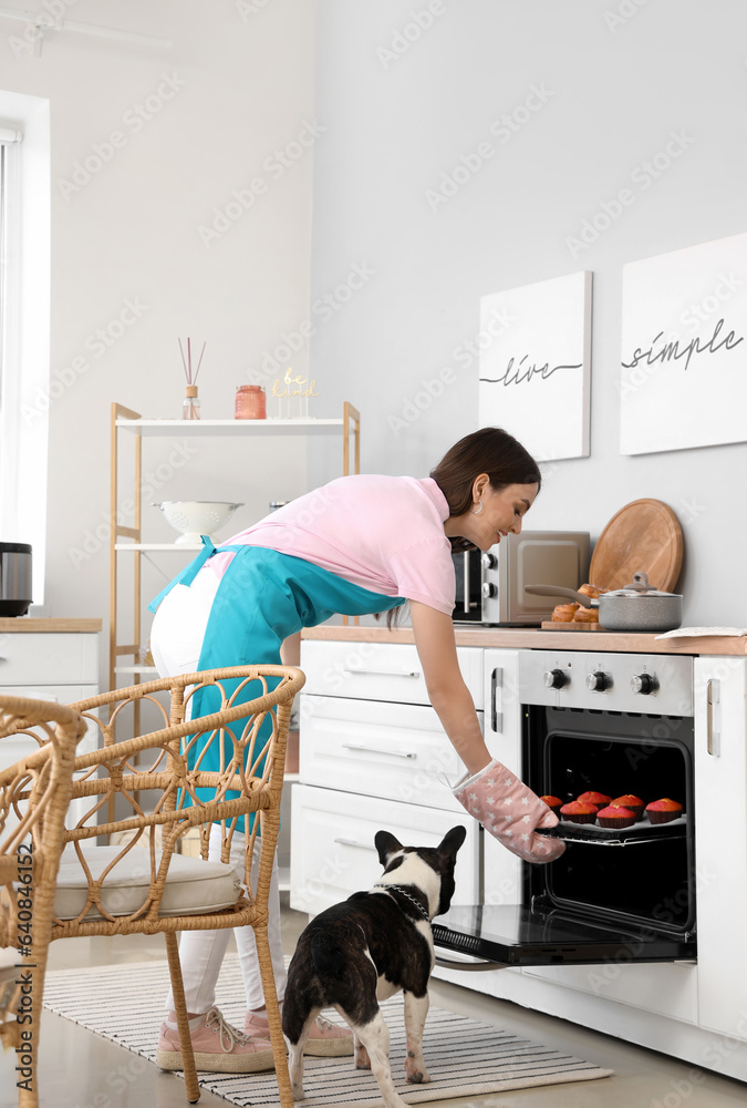 Young woman taking tray with muffins from electric oven in kitchen