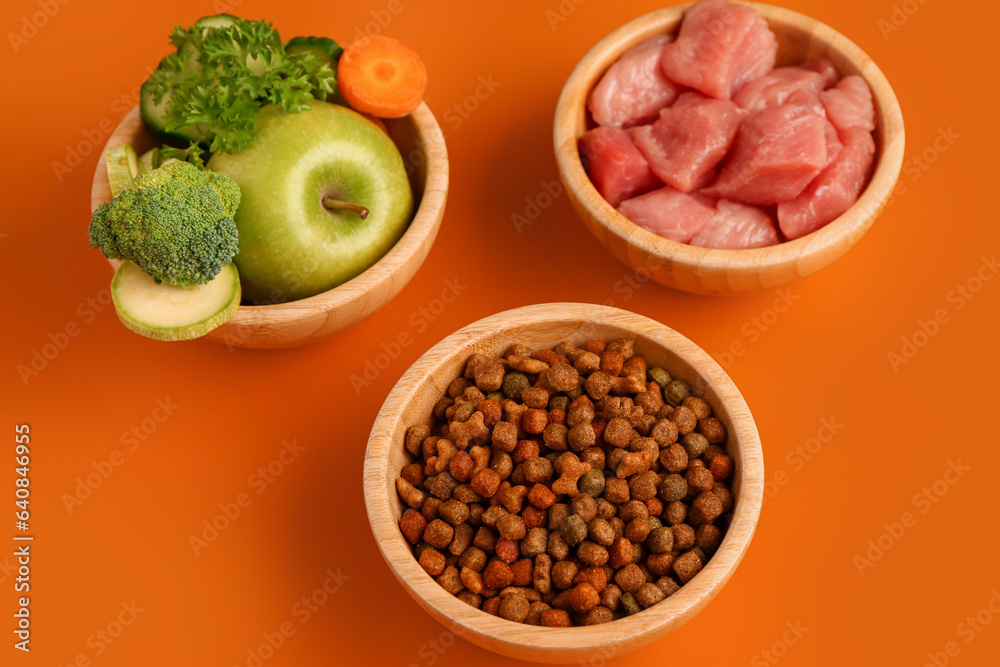 Bowls with dry pet food, raw meat and natural products on orange background