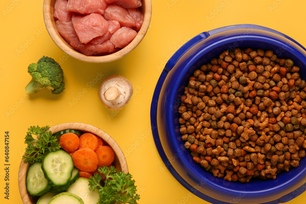 Bowls with dry pet food, raw meat and natural products on yellow background