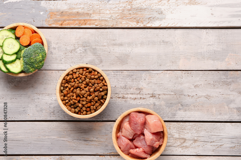 Bowls with dry pet food, raw meat and natural products on light wooden background