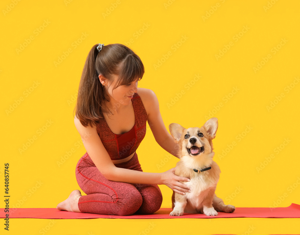 Sporty young woman sitting on yoga mat with cute Corgi dog against yellow background