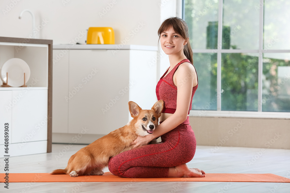 Sporty young woman and cute Corgi dog sitting on yoga mat in kitchen