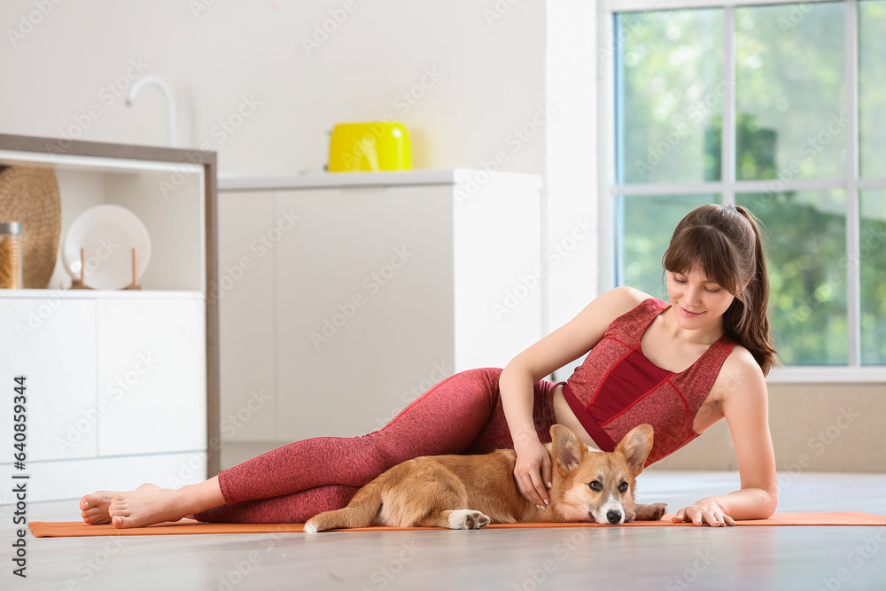 Sporty young woman and cute Corgi dog lying on yoga mat in kitchen