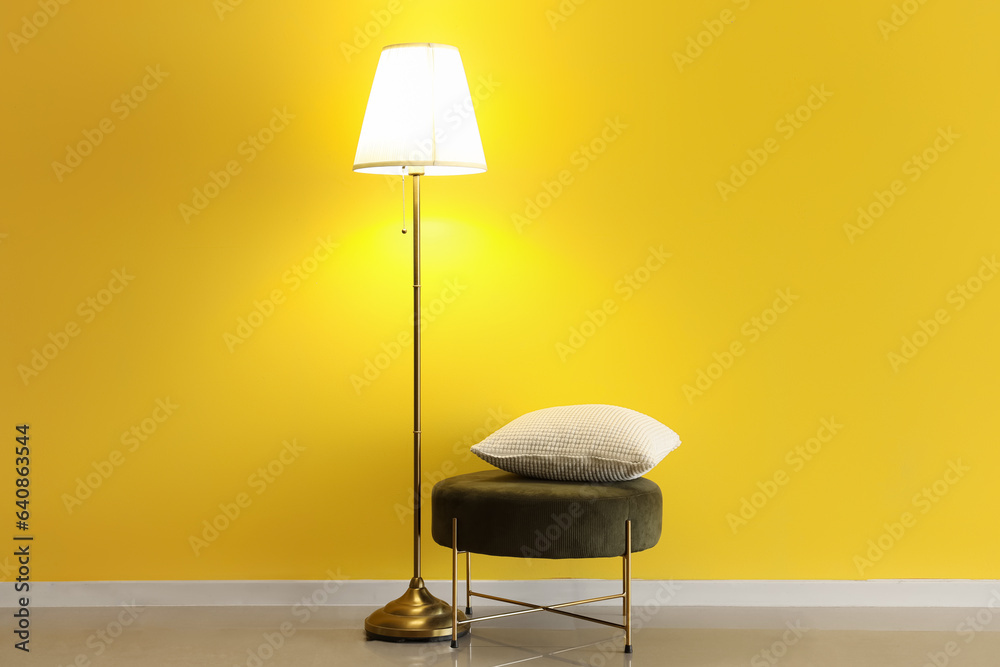 Glowing stand lamp and pouf with pillow near yellow wall in room