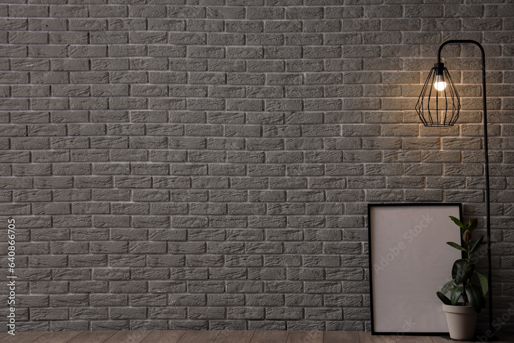 Glowing lamp, houseplant and blank frame near grey brick wall