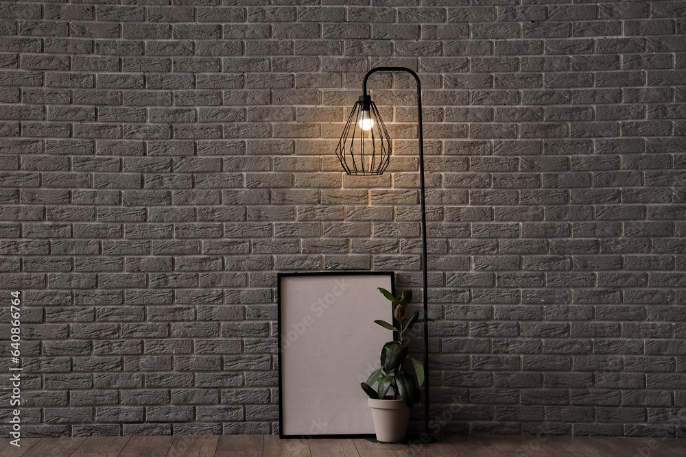 Glowing lamp, houseplant and blank frame near grey brick wall