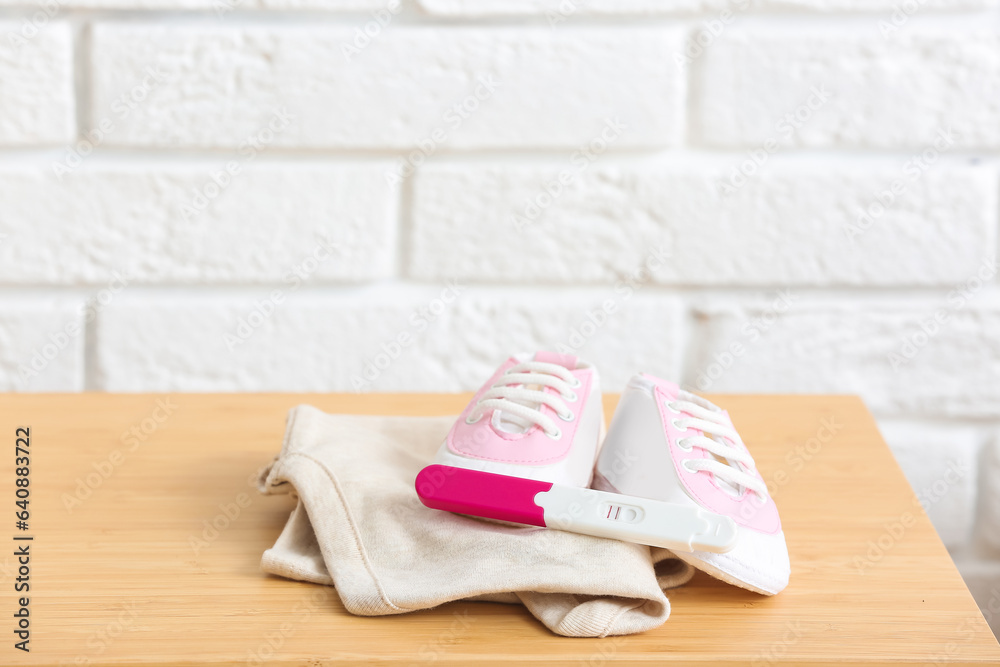 Baby booties, clothes and pregnancy test on wooden table against light brick wall