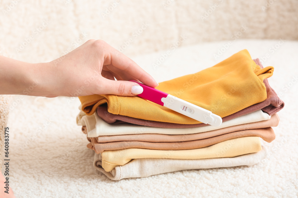 Female hand with pregnancy test and stack of baby clothes on sofa