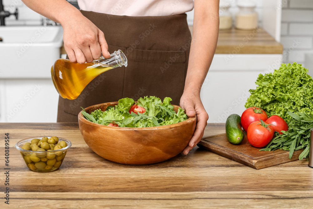 Woman adding olive oil into bowl with tasty salad at table in kitchen