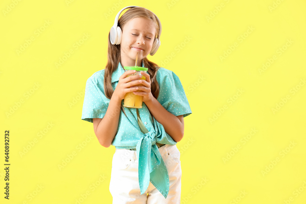 Little girl in headphones with glass of fresh citrus juice on yellow background