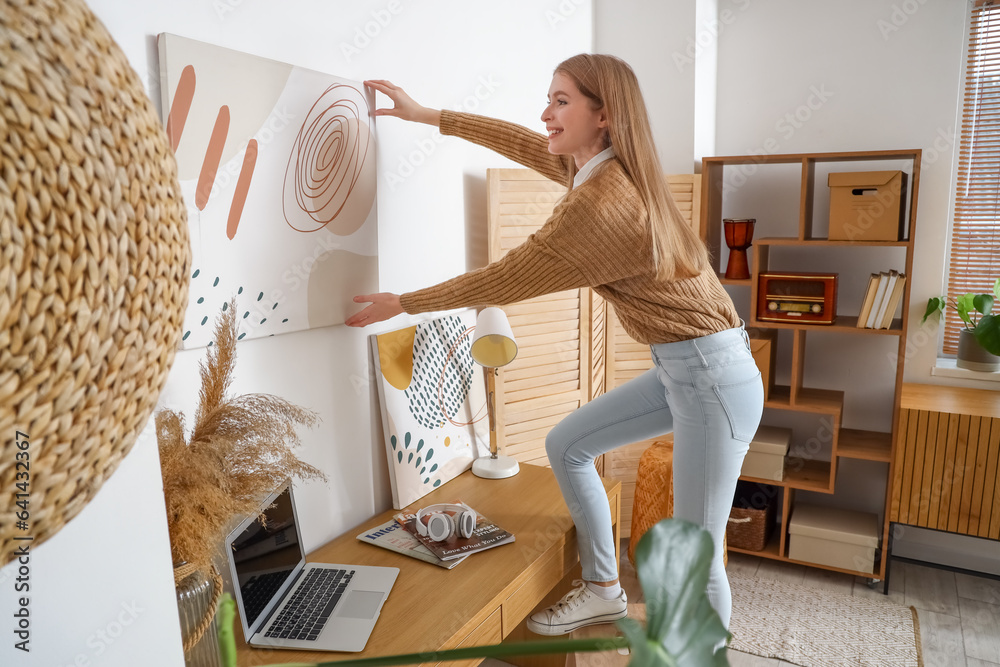 Young woman hanging painting on light wall at home