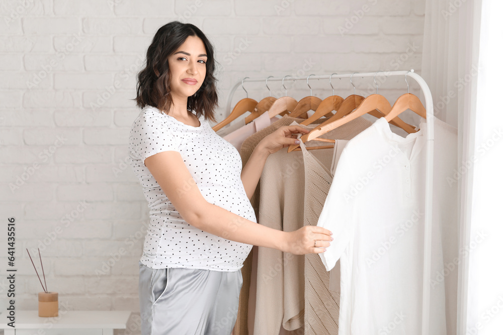 Young pregnant woman near rack with clothes in bedroom