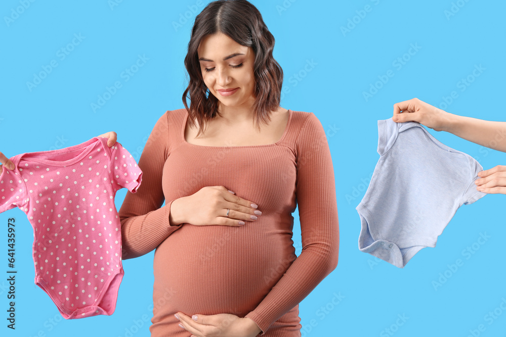 Young pregnant woman and female hands with baby bodysuits on blue background