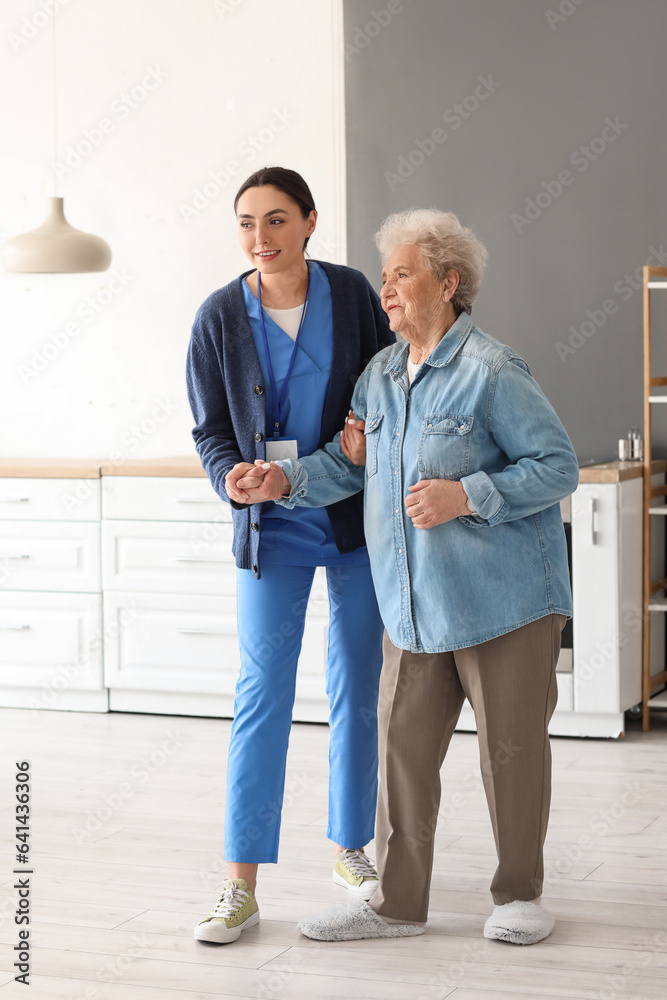 Young caregiver helping senior woman to walk in kitchen