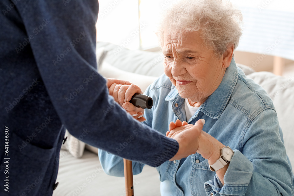 Young caregiver helping senior woman with stick to stand up at home, closeup