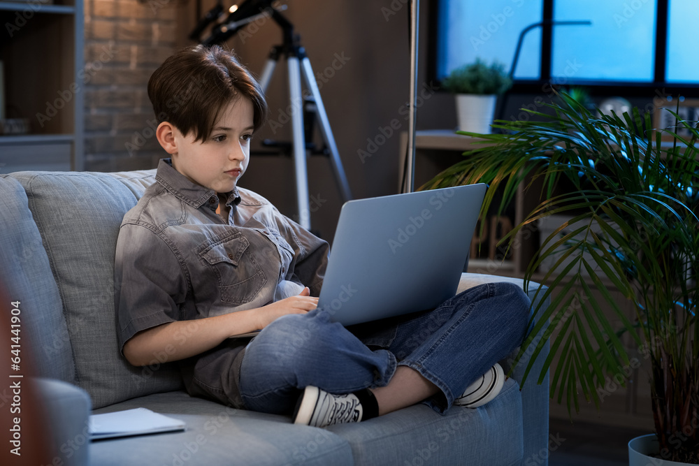 Little boy doing homework with laptop and sitting on sofa at home in evening