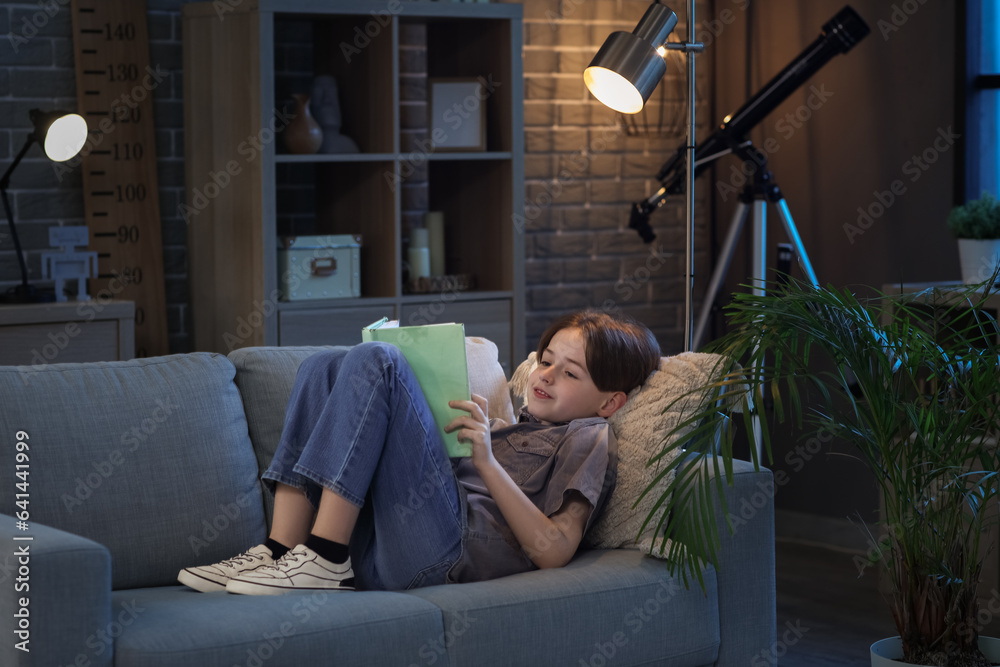 Little boy reading book and lying on sofa at home in evening