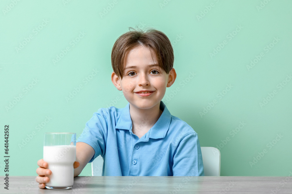 Happy little boy sitting at table with glass of fresh milk on turquoise background