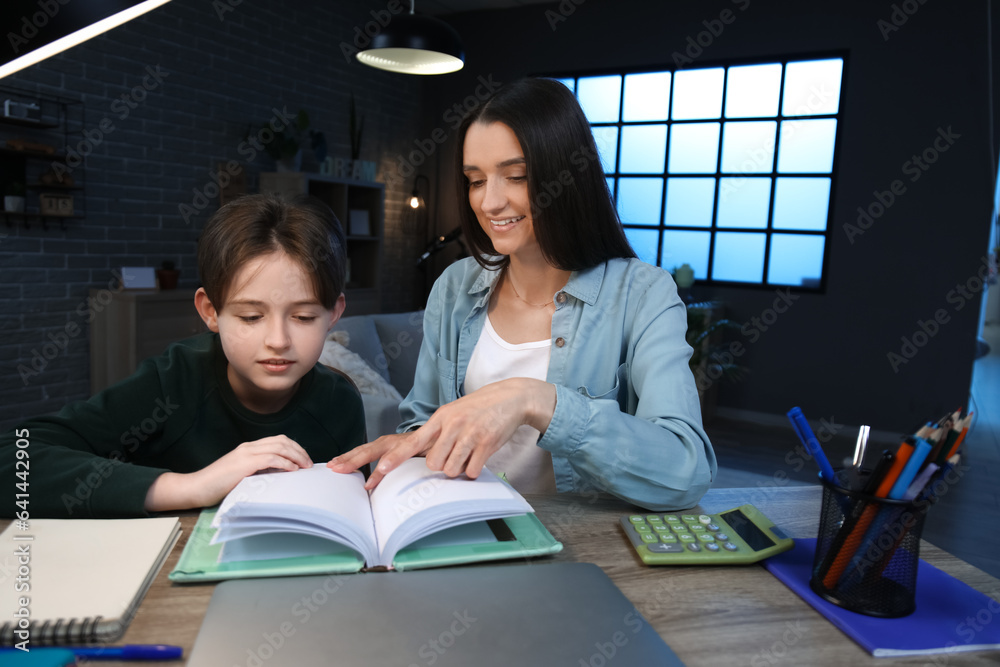 Mother with her little son sitting at table and reading book late in evening