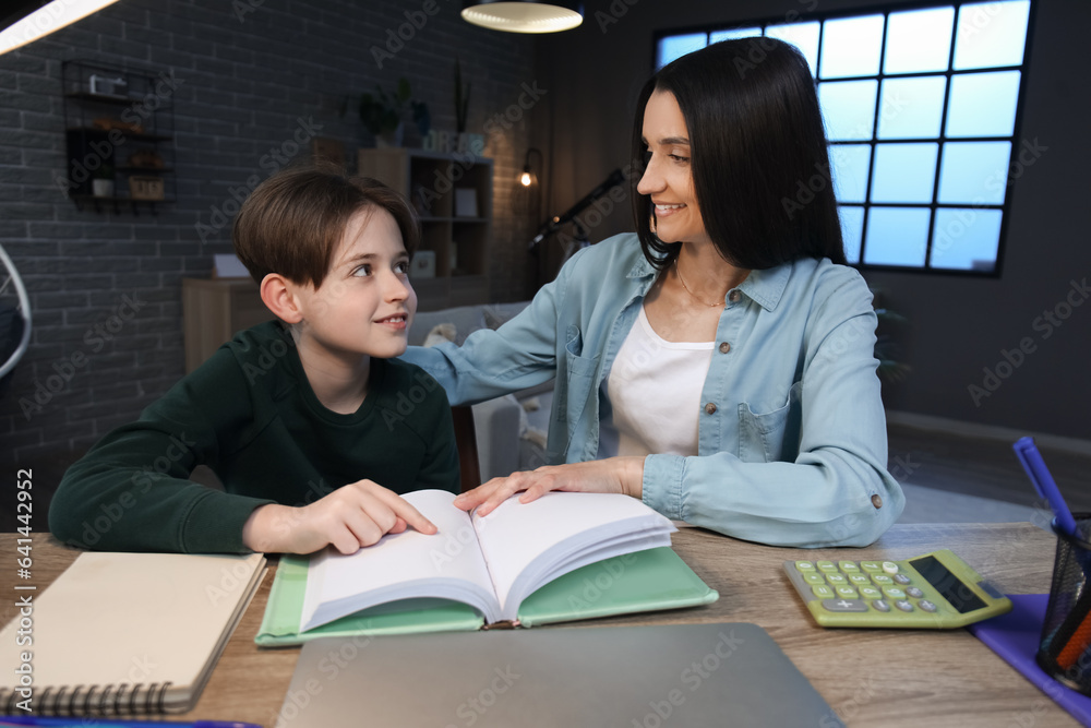 Mother with her little son sitting at table and reading book late in evening