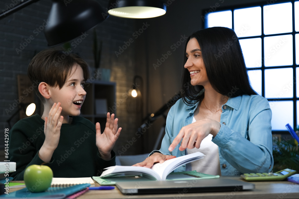 Mother with her little son sitting at table and reading book late in evening