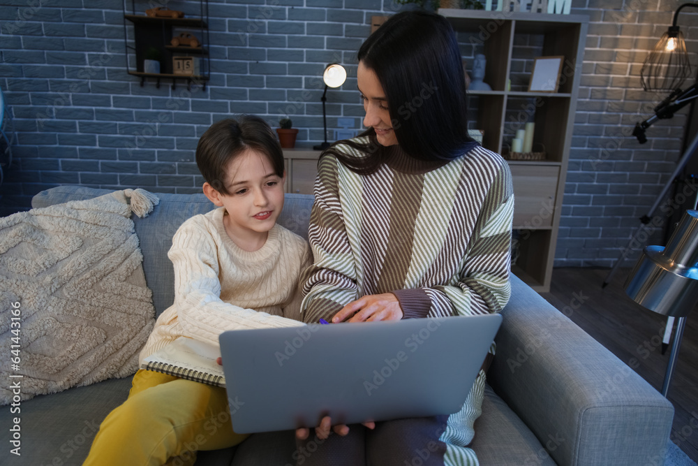 Little boy and his mother doing homework with laptop at home late in evening