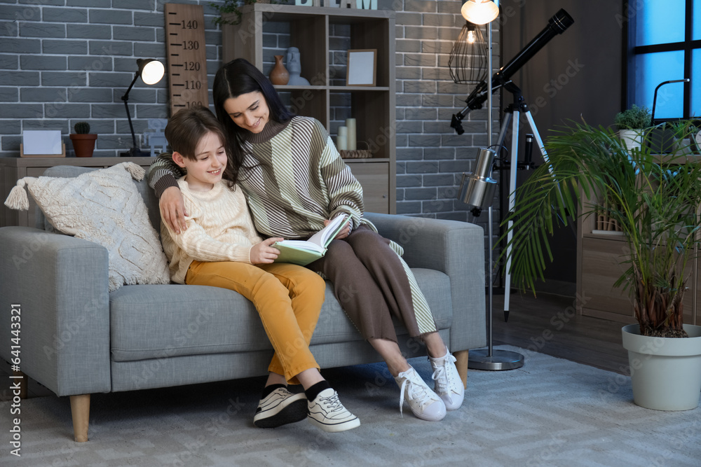 Little boy with his mother sitting on sofa and reading book at home late in evening